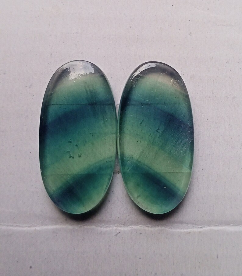 Puede incluir: Dos cabujones ovalados de fluorita verde y azul con un degradado de color verde claro y blanco. Los cabujones son lisos y pulidos.