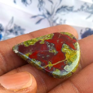 Puede incluir: Un cabuj&oacute;n de &aacute;gata rojo, verde y marr&oacute;n con un patr&oacute;n &uacute;nico. El cabuj&oacute;n tiene forma de l&aacute;grima y una superficie lisa y pulida.