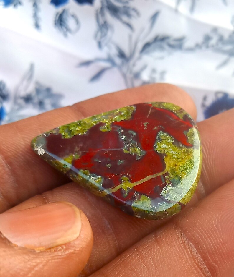 Puede incluir: Un cabuj&oacute;n de &aacute;gata rojo, verde y marr&oacute;n con un patr&oacute;n &uacute;nico. El cabuj&oacute;n tiene forma de l&aacute;grima y una superficie lisa y pulida.