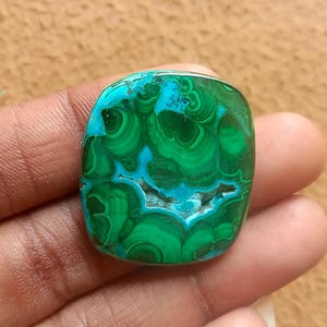 May include: A green and blue malachite cabochon gemstone with a swirling pattern. The stone is cut in a rectangular shape and has a smooth, polished surface.
