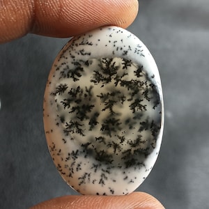 May include: Oval gemstone featuring a white background with black, tree-like dendritic inclusions. The polished stone is held between fingers, highlighting its natural patterns. A unique piece of jewellery, the stone's measurements are in centimetres.