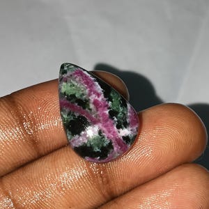 Puede incluir: Una piedra preciosa pulida con forma de lágrima. La piedra presenta una mezcla de colores, incluyendo rosa intenso, verde, negro y blanco. La gema se sostiene en una mano, mostrando su belleza natural y patrones de color únicos.