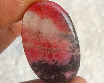 Könnte beinhalten: Oval geformter Edelstein mit polierter Oberfläche. Der Stein zeigt eine Mischung aus Farben, darunter Rot, Weiß, Rosa und Schwarz. Der Edelstein wird zwischen Fingern gehalten und zeigt seine natürliche Schönheit und einzigartigen Farbvariationen.