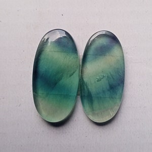 Puede incluir: Dos piedras preciosas de fluorita ovaladas de color verde y azul con un acabado liso y pulido.