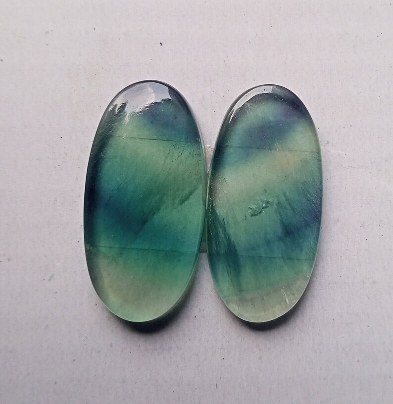 Puede incluir: Dos piedras preciosas de fluorita ovaladas de color verde y azul con un acabado liso y pulido.