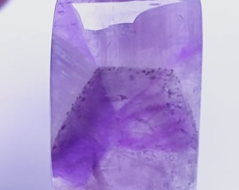 May include: A rectangular amethyst gemstone with a smooth, polished surface. The stone displays a translucent light purple hue with darker violet inclusions forming a distinct geometric pattern within the center of the crystal structure.