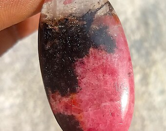 Könnte beinhalten: Oval geformter Edelstein mit polierter Oberfläche. Der Stein weist eine Mischung aus Farben auf, darunter ein leuchtendes Rosa, tiefes Schwarz und ein durchscheinender weißer Bereich. Der Edelstein wird zwischen den Fingern gehalten, um seine Größe und Form zu zeigen.