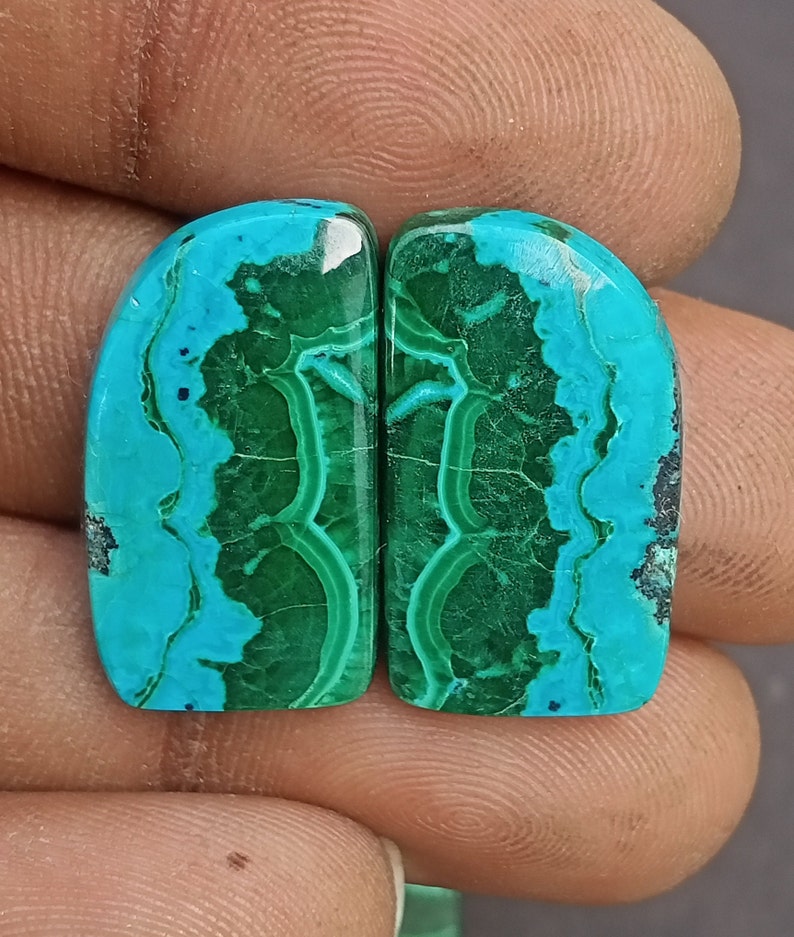 May include: Two green and blue gemstone cabochons with a marbled pattern. The cabochons are cut in a rounded rectangle shape.
