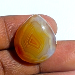 May include: A teardrop-shaped agate cabochon with a banded pattern of orange, yellow, and white. The stone is translucent and has a glossy finish.