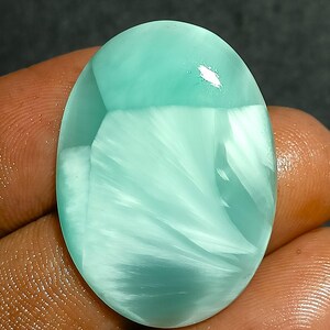 Puede incluir: Gema ovalada pulida, de color verde azulado claro, con un patrón blanco y ondulado. La gema se sostiene en una mano, mostrando su superficie lisa y reflectante. El diseño se asemeja a una nube o una ola.