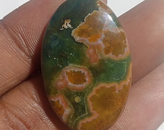 May include: An oval-shaped gemstone with a polished surface. The stone features a mix of colors, including green, brown, and orange, with a unique pattern of swirls and spots. The gemstone is held in a hand.