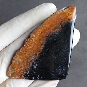 May include: A polished, triangular gemstone with a striking contrast of colors. The top edge features a band of orange and brown hues, transitioning to a deep black base. Small white flecks are visible.