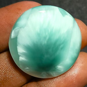 Puede incluir: Una gema redonda y pulida con un patrón arremolinado de tonos blancos y verde-azules. La piedra tiene una superficie lisa y brillante y se sostiene en una mano. El color de la gema se asemeja a las olas del océano. Esta gema probablemente se usa en joyería.