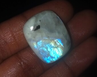 May include: A polished, cabochon-cut moonstone gemstone. The stone has a translucent, milky white base with a flash of blue and gold iridescence. A small black inclusion is visible near the top. The gemstone is held in a hand.