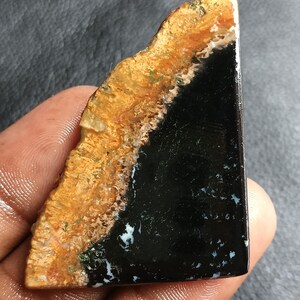May include: A polished, triangular-shaped gemstone cabochon featuring a distinct color split. One side displays a textured, golden-brown, and tan earthy pattern, while the other side is a smooth, deep black surface with small, scattered white inclusions.
