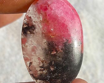 Puede incluir: Una gema ovalada con un llamativo degradado de color. La mitad superior es de un rosa vibrante, que transiciona a blanco y negro en la parte inferior. La piedra se sostiene entre dos dedos, mostrando su superficie pulida.