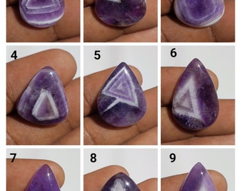 Könnte beinhalten: Neun Amethyst-Cabochons in verschiedenen Formen, darunter Tropfen- und Herzformen, mit einem weißen, dreieckigen Muster im Inneren. Die Cabochons werden auf einer Hand gezeigt.