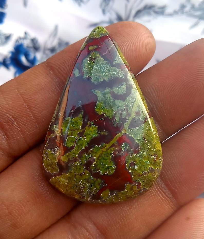 K&ouml;nnte beinhalten: Ein tropfenf&ouml;rmiger Cabochon aus gr&uuml;nem, rotem und braunem Jaspis. Der Stein hat ein geflecktes, wirbelndes Muster.