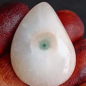 Può includere: Una gemma lucida a forma di goccia, con una base bianca e un vortice centrale verde e marrone. La pietra è tenuta in mano, evidenziando la sua superficie liscia e riflettente. Il motivo unico della gemma la rende un pezzo sorprendente.