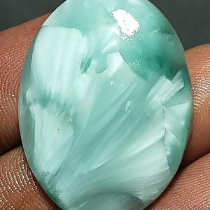 Può includere: Pietra preziosa ovale con un motivo a spirale di bianco e verde acqua chiaro. La superficie lucida riflette la luce, evidenziando le trame naturali e le variazioni di colore della pietra. La gemma è tenuta in una mano.