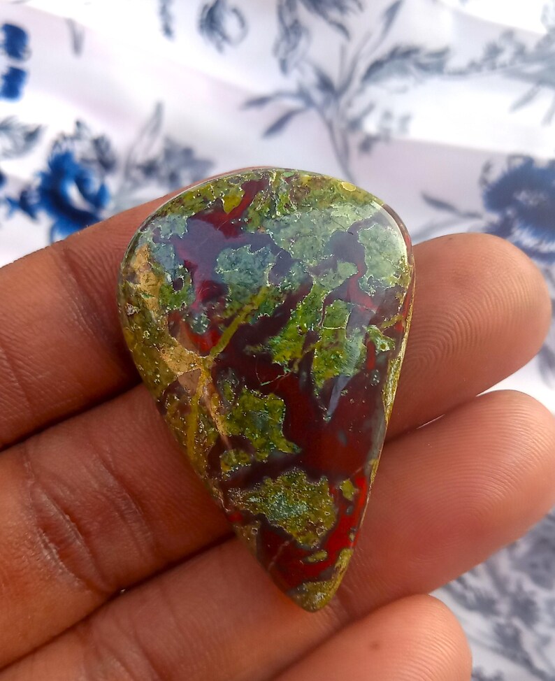 K&ouml;nnte beinhalten: Ein tropfenf&ouml;rmiger Cabochon aus gr&uuml;nem, rotem und braunem Jaspis. Der Stein hat ein einzigartiges Muster aus wirbelnden Farben und Texturen.