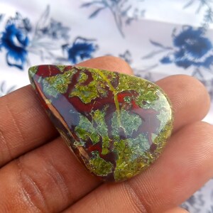 K&ouml;nnte beinhalten: Ein gr&uuml;ner, roter und brauner Jaspis-Cabochon in Tropfenform. Der Stein hat ein einzigartiges Muster aus wirbelnden Farben und Texturen.