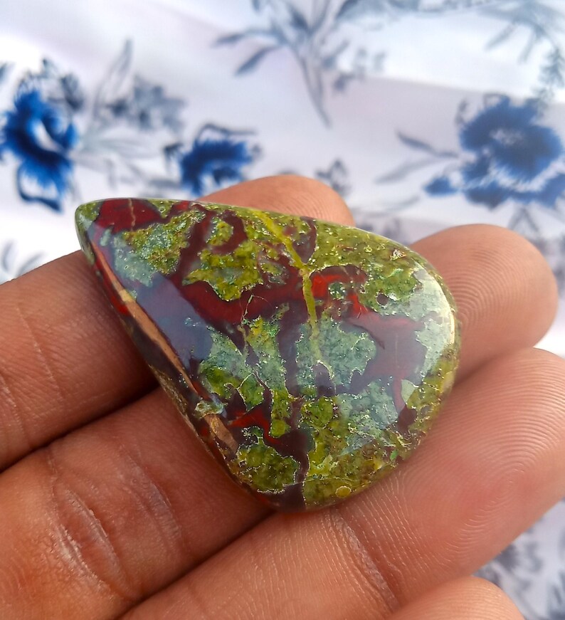 K&ouml;nnte beinhalten: Ein gr&uuml;ner, roter und brauner Jaspis-Cabochon in Tropfenform. Der Stein hat ein einzigartiges Muster aus wirbelnden Farben und Texturen.
