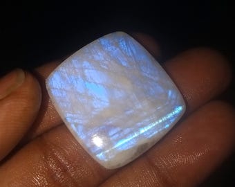 May include: A square-cut, iridescent gemstone with a pearly white base and blue internal glow. The stone is held in a hand, showcasing its polished surface and natural patterns. The gem's corners are slightly rounded.