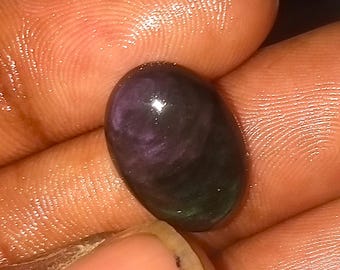 May include: An oval-shaped gemstone with a polished surface. The stone displays a blend of dark purple, green, and black hues. The gemstone is held in a hand, showcasing its size and color variations.
