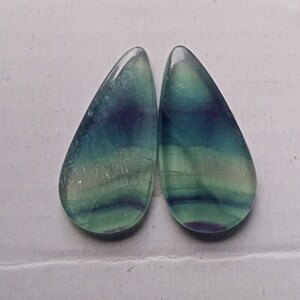 Puede incluir: Par de cabujones de fluorita en forma de l&aacute;grima, de color verde, azul y morado. Los cabujones son transl&uacute;cidos y tienen un patr&oacute;n estriado.