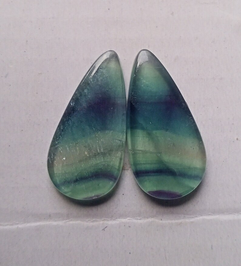 Puede incluir: Par de cabujones de fluorita en forma de l&aacute;grima, de color verde, azul y morado. Los cabujones son transl&uacute;cidos y tienen un patr&oacute;n estriado.