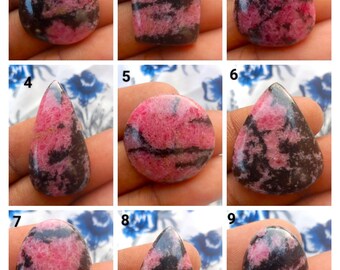 Könnte beinhalten: Neun polierte Rhodonit-Edelsteine in verschiedenen Formen. Die Steine zeigen eine Mischung aus rosa und schwarzen Mustern. Zu den Formen gehören Tropfen, Rechteck, Oval und Rund. Die Edelsteine werden vor einem neutralen Hintergrund präsentiert.