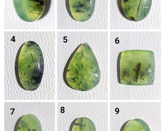 May include: Nine prehnite gemstones in various shapes, including oval, teardrop, and square. The translucent stones are a pale green color with dark green and black inclusions. The gemstones are displayed against a white background.