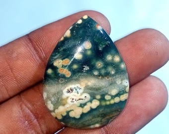 Puede incluir: Un cabujón de jaspe oceánico verde, blanco y marrón con forma de lágrima. La piedra tiene un patrón único de manchas y remolinos blancos y marrones.