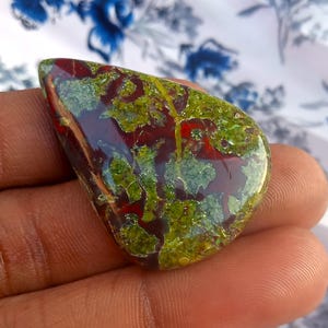 K&ouml;nnte beinhalten: Ein gr&uuml;ner, roter und brauner Cabochon-Edelstein mit einem einzigartigen, marmorierten Muster. Der Stein ist poliert und hat eine glatte, gl&auml;nzende Oberfl&auml;che.
