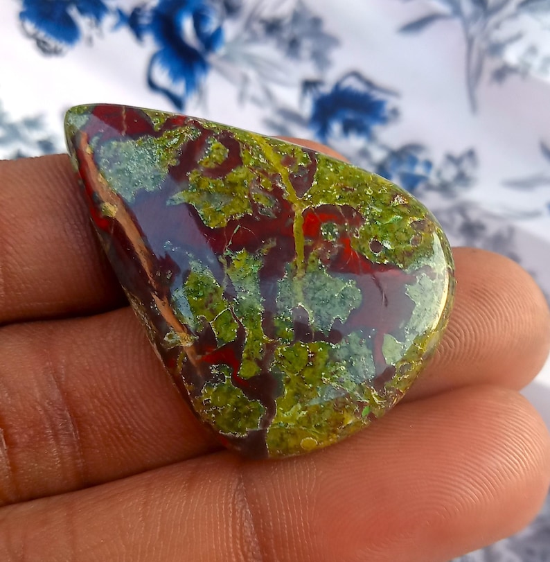 K&ouml;nnte beinhalten: Ein gr&uuml;ner, roter und brauner Cabochon-Edelstein mit einem einzigartigen, marmorierten Muster. Der Stein ist poliert und hat eine glatte, gl&auml;nzende Oberfl&auml;che.