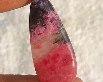 Könnte beinhalten: Ein tropfenförmiger, polierter Edelstein. Der Stein zeigt eine Mischung aus Farben, darunter tiefes Rot, Rosa, Schwarz und Weiß. Der Edelstein wird zwischen Fingern gehalten, was seine Form hervorhebt. Dies ist ein natürlicher Edelstein.