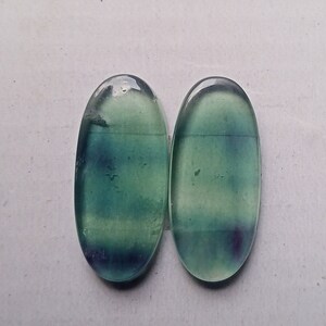 Puede incluir: Dos cabujones ovalados de fluorita verde y azul. Las piedras tienen una apariencia transl&uacute;cida con un ligero brillo.