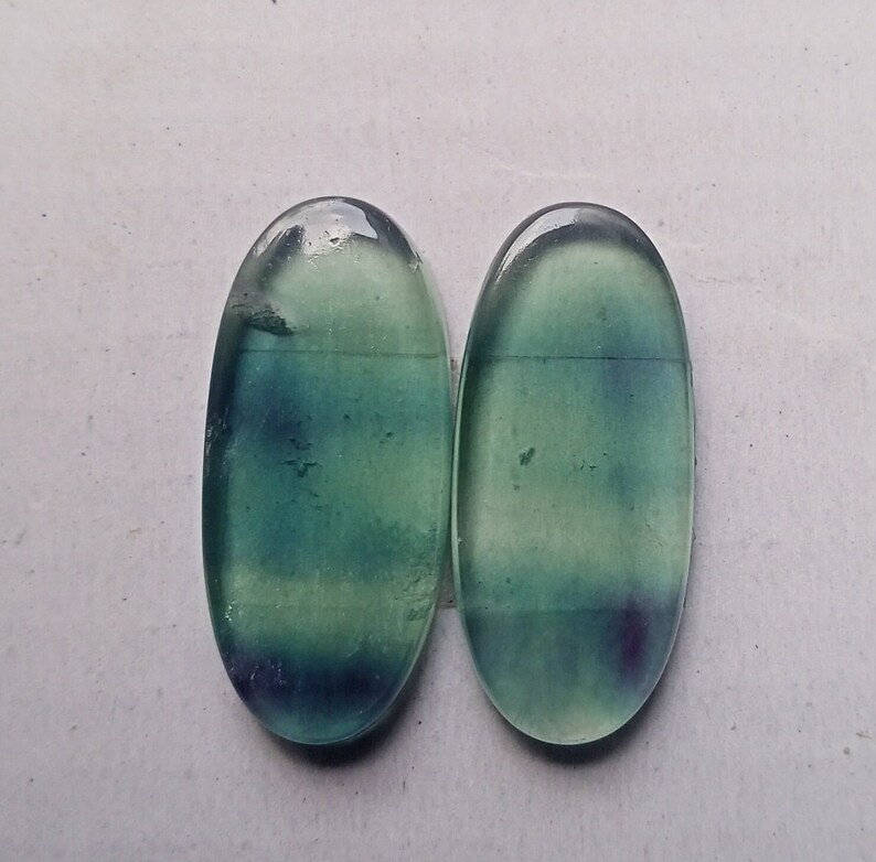 Puede incluir: Dos cabujones ovalados de fluorita verde y azul. Las piedras tienen una apariencia transl&uacute;cida con un ligero brillo.