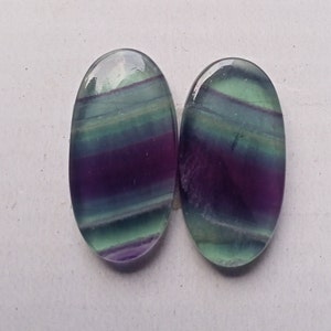 Puede incluir: Dos cabujones ovalados de fluorita, de color verde, morado y azul. Los cabujones tienen un patr&oacute;n rayado.