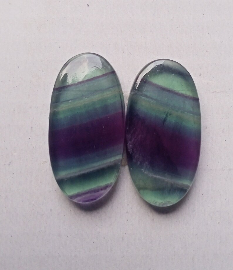 Puede incluir: Dos cabujones ovalados de fluorita, de color verde, morado y azul. Los cabujones tienen un patr&oacute;n rayado.