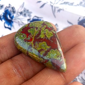 K&ouml;nnte beinhalten: Ein gr&uuml;ner, roter und brauner Edelstein-Cabochon in Tropfenform. Der Stein hat ein einzigartiges Muster aus wirbelnden Farben und Texturen.
