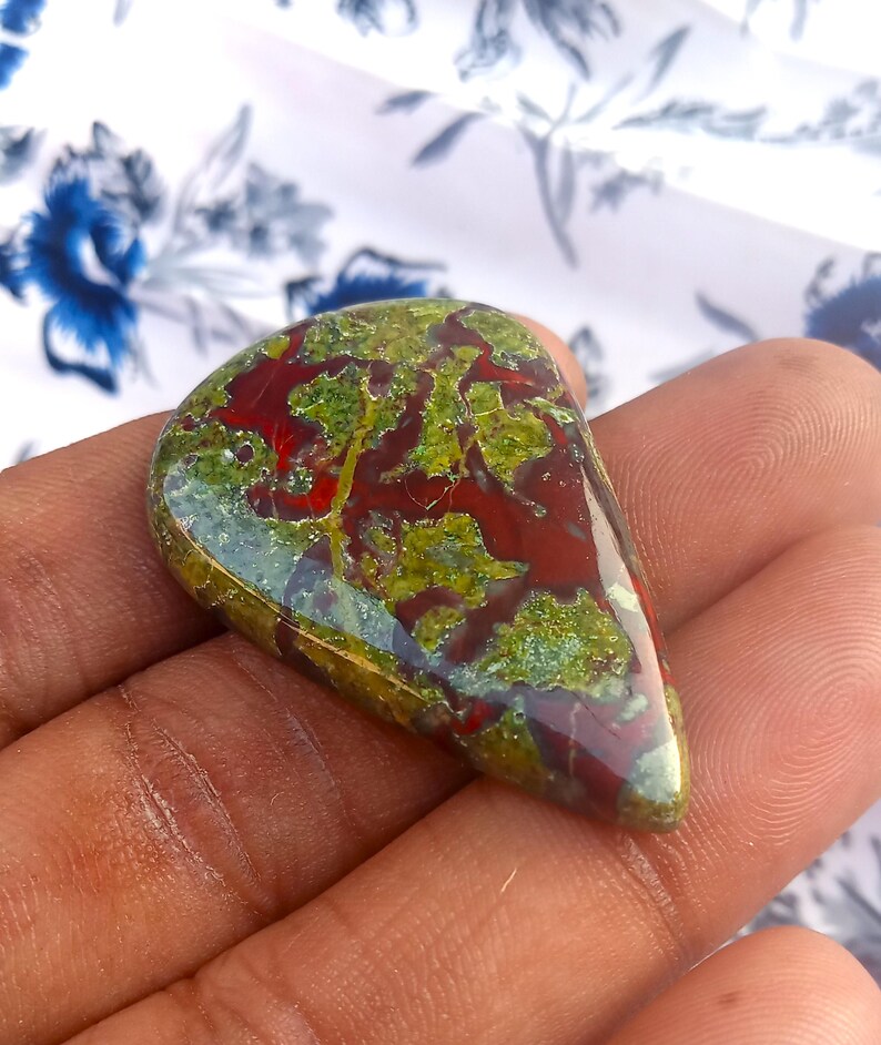 K&ouml;nnte beinhalten: Ein gr&uuml;ner, roter und brauner Edelstein-Cabochon in Tropfenform. Der Stein hat ein einzigartiges Muster aus wirbelnden Farben und Texturen.