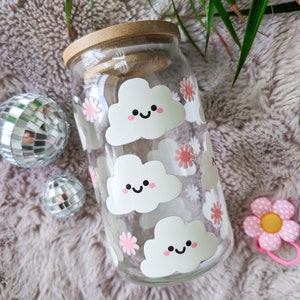 May include: A clear glass jar with a bamboo lid, decorated with white clouds with pink flowers and smiling faces. The jar is sitting on a fluffy white surface.