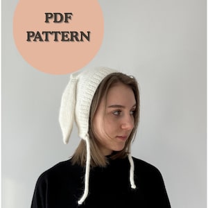 Peut inclure: Un bonnet de lapin blanc tricoté avec de longues oreilles souples et des liens attachés. Le chapeau est fait d'un fil texturé. L'image comprend un cercle pêche avec le texte "PDF PATTERN".