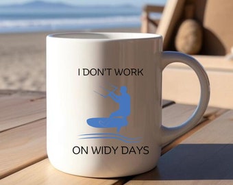 May include: White ceramic mug featuring a blue kite surfer graphic and the text "I DON'T WORK ON WIDY DAYS". The mug is on a wooden table with a beach and sea in the background.
