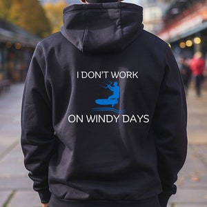 Peut inclure: Sweat-shirt à capuche noir avec le texte "Je ne travaille pas les jours de vent" et un graphique d'une personne en kitesurf.