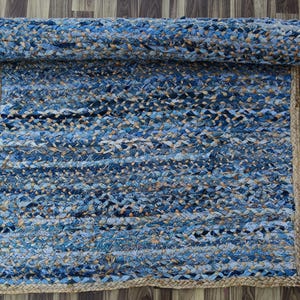May include: A rolled-up blue and tan braided rug with a jute border. The rug is made of woven strips of denim in varying shades of blue and tan. The rug is rectangular and has a natural jute border.