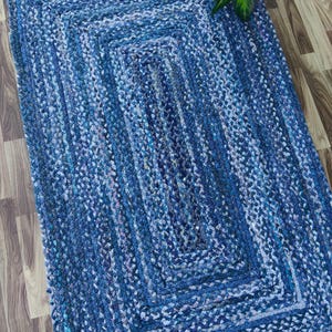 Puede incluir: Una alfombra rectangular trenzada en tonos azules, creando una superficie texturizada. La alfombra presenta un diseño rectangular anidado, con variaciones de azul y blanco. La alfombra mide aproximadamente 91 cm de ancho y 152 cm de largo.