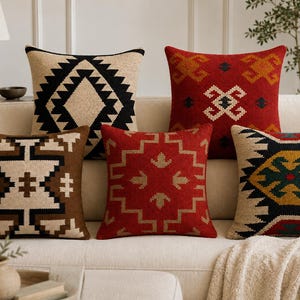 May include: Five square throw pillows featuring woven geometric patterns in various color combinations including black and beige, solid red with white and orange accents, brown and beige, red with beige, and a multi-colored design with green and yellow.